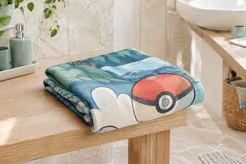 Serviette de plage Pokémon pliée sur un banc en bois blond dans une salle de bain lumineuse.