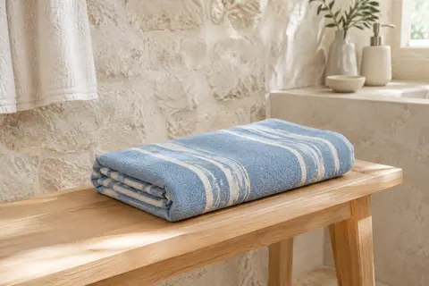 Serviette de plage bleu ciel à rayures, pliée sur un banc en bois clair dans une salle de bain lumineuse.