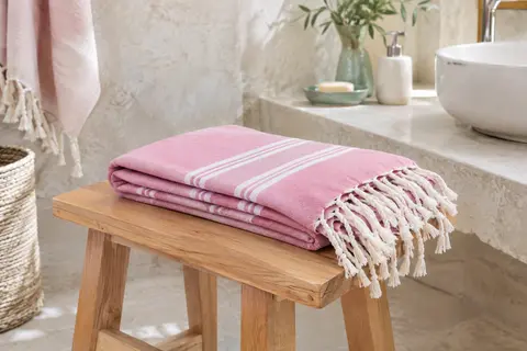 Fouta de plage rose à rayures et franges pliée sur un banc en bois clair dans une salle de bain lumineuse