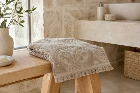 Serviette de hammam beige en coton jacquard baroque pliée sur un banc de bois clair dans une salle de bain minérale.