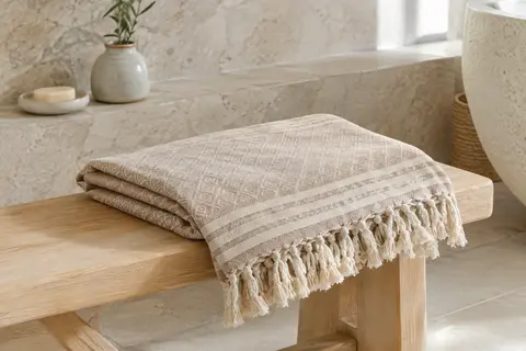 Fouta de plage en coton sable à motif rayé, pliée sur un banc en bois clair dans une salle de bain lumineuse.