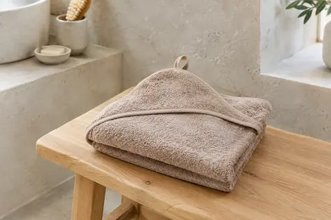 Cape de bain bébé en éponge de coton nougat, pliée avec sa capuche visible dans une salle de bain lumineuse