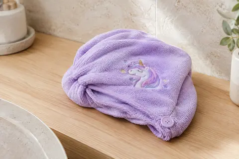 Turban pour cheveux lilas à motif licorne, plié sur une tablette en bois blond dans une salle de bain claire.