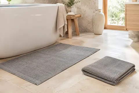 Deux tapis de bain gris en coton, l’un posé au sol et l’autre plié dans une salle de bain claire.