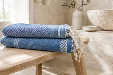 Deux foutas de plage en coton bleu à motif graphique, pliées sur un banc en bois clair dans une salle de bain lumineuse.